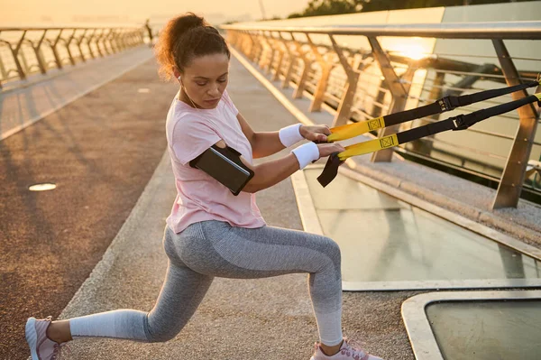 Athletic sportswoman exercises with suspension straps during body weight training at the urban environment of a city bridge. Full body female athlete in sportswear pulling suspension ropes and lunging