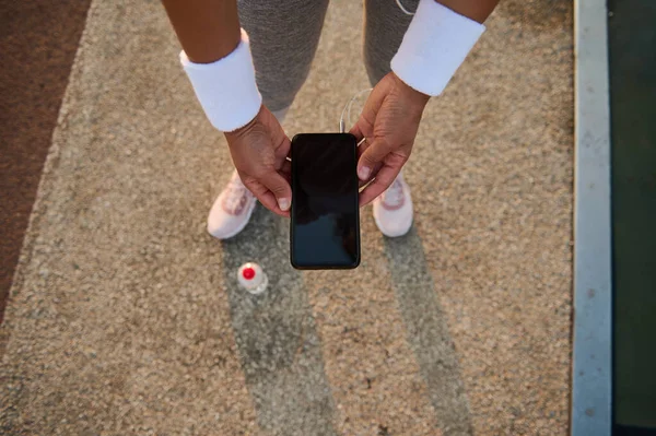High angle view of female athlete hands in white terry wristbands holding a smartphone with empty blank black screen and browsing on websites and mobile applications.