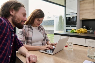 Dizüstü bilgisayarda çalışan, klavyeye yazı yazan kocasının yanında mutfak masasında oturan monitör ekranına bakıp gülümseyen bir kadın. Çevrimiçi alışveriş ve uzak çalışma kavramı
