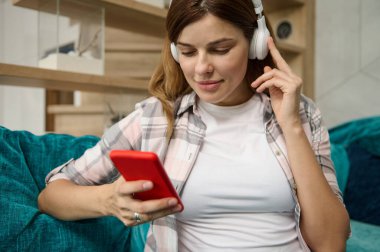 Kafkas asıllı hamile bir kadının portresi. Kulaklığı akıllı telefonuyla, internet sitelerini ve mobil uygulamaları kullanarak evdeki kanepede otururken dinleniyor.