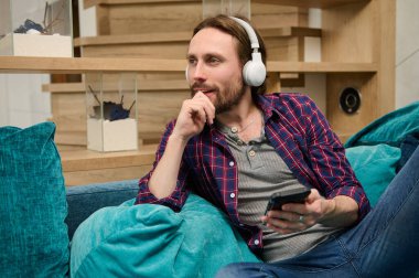 Orta yaşlı, sakallı, kablosuz kulaklıklı yakışıklı beyaz adam elinde akıllı bir telefon tutuyor ve rüya gibi bir şekilde yan tarafa bakıyor, izin gününün tadını çıkarıyor, ev ortamındaki bir kanepede dinleniyor.