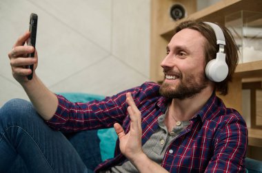 Yakışıklı adam video konuşmalarıyla eğleniyor, kablosuz kulaklık takıyor ve gülümsüyor akıllı telefon web kamerasına bakıyor, selamlıyor ve evde dinlenirken el sallıyor.