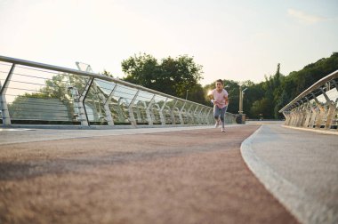 Tam boy. Kararlı kadın atlet koşu bandı üzerinde köprü boyunca koşar, şafakta sabah kardiyo antrenmanı yapar. Açık hava sporları eğitimi, kilo kaybı, spor, spor, aktif yaşam tarzı konsepti