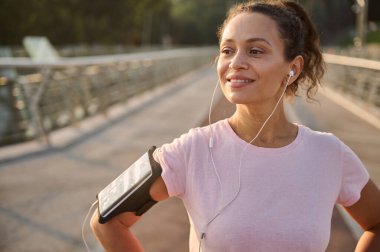 Mutlu bir İspanyol spor kıyafetinin mutlu bir portresi, akıllı telefon kılıfı ve kulaklık takıyor, gülümsüyor, köprüde duruyor, koşudan sonra dinleniyor.