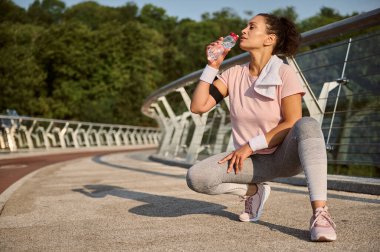 Güzel İspanyol sporcu kadın pembe tişört ve gri içme suyu giyer şehir köprüsünde çömelmiş bir pozisyonda oturur ve güneş doğarken sabah koşusu ve kardiyo antrenmanından sonra dinlenir.