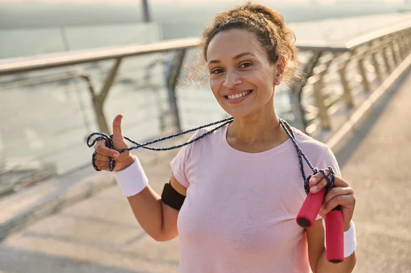 Happy Hispanic sportswoman with a skipping rope stands on the modern glass city bridge and smiles with beautiful and cheerful toothy smile looking at camera, enjoying morning cardio workout outdoor