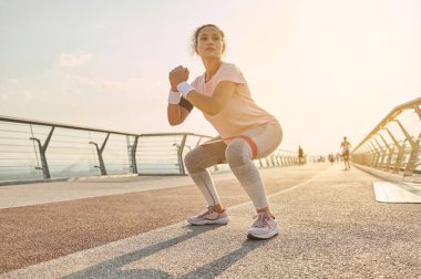 Kararlı orta yaşlı sporcu kadın, gün doğumunda dışarıda egzersiz yapan bayan sporcu elastik direniş bandıyla çömelme hareketleri yapıyor. Fitness spor dayanıklılığı, vücut ağırlığı eğitimi ve aktif yaşam tarzı konsepti