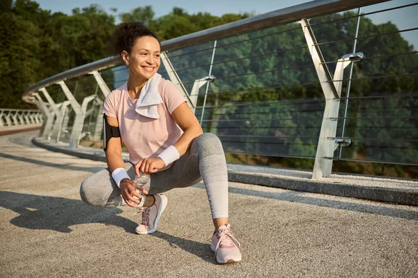 Portrait of a female athlete in sportswear and white terry wristbands holding a bottle of water sitting on squat pose on the treadmill, smiles looking away relaxing after morning jog along the bridge