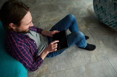 Kafkas sakallı, sıradan kıyafetler giyen genç bir adamın yukarıdan görünüşü evde dijital tabletle çalışıyor. Günlük hayatta dijital elektronik cihazlar var. İnternet, çevrimiçi, uzak çalışma, bağlantı kavramı