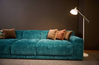 Mobilya mağazasının galerisinde modern şık döşenmiş mobilya sergisi. Bir turkuaz yumuşak kadife kanepeye odaklan ve bir lamba tarafından aydınlatılan kahverengi yastıklar kahverengi bir duvar arka planına.