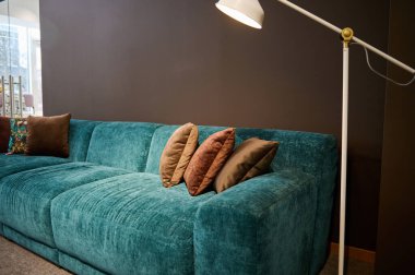 Mobilya mağazasının galerisinde modern şık döşenmiş mobilya sergisi. Bir turkuaz yumuşak kadife kanepenin yakın çekimi ve bir lamba tarafından yakılan kahverengi yastıklar kahverengi bir duvar arka planına karşı