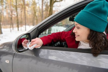 Genç bir kadın sürücü arabasının şoför koltuğunda oturur ve güzel, soğuk bir kış günü karlı bir havada seyahat ederken karla kaplı bir orman sahasında dururken sürücü tarafındaki aynayı siler.