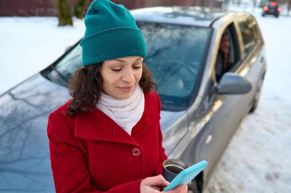 Очаровательная женщина, водитель путешественник сидит на капоте автомобиля в заснеженном лесу, держит мобильный телефон во время отдыха во время ее поездки на машине, наслаждаясь путешествием в заснеженной природе в зимний прохладный день