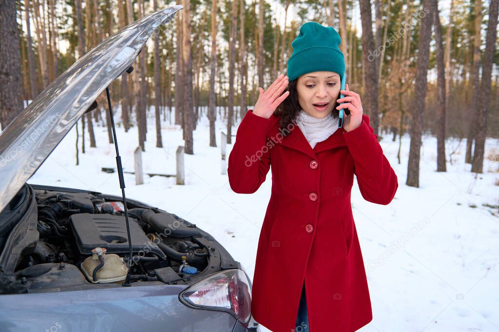 Shocked frustrated middle aged woman standing near her broken car and trying to call for roadside assistance during a car breakdown on a snowy road. Auto insurance and travel concepts