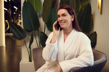 Modern lüks spa kliniğinde güzellik ve güzellik tedavisi gördükten sonra beyaz atlı beyaz bornozlu çekici bir kadın cep telefonuyla spa salonunda oturuyor.
