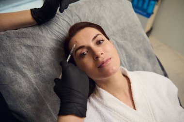 Yüksek açılı kozmetik uzmanı elinde botulinyum toksin ya da hiyalüronik asit olan bir şırınga tutan ve sağlık merkezindeki orta yaşlı beyaz kadının yüzüne iğne yapan tıbbi eldivenler verdi.