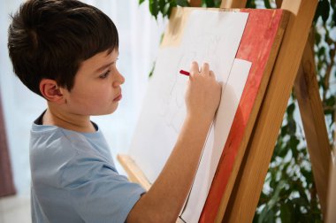 Yakışıklı, konsantre olmuş beyaz okul çocuğu evdeki ahşap bir sehpaya tuval çiziyor. Çocuk eğlencesi, çocuk sanat sınıfı, hobi, eğitim ve yaratıcılık kavramı