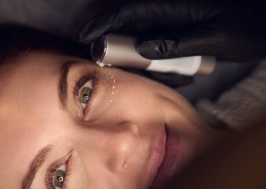 Güzel, orta yaşlı, esmer, beyaz bir kadının gözlerinin altına bir damla kozmetik ürün veya serum damlatan portresi. Sağlık merkezinden profesyonel cilt bakımı alıyor.