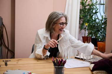 Zarif, neşeli Avrupalı yaşlı kadın, yaratıcı atölyede kadın moda tasarımcısı, çalışırken ahşap masada oturuyor ve giysiler için çizimler yapıyor..