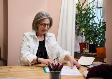 Zarif, neşeli, olgun Kafkasyalı kadın, yaratıcı atölyede kadın moda tasarımcısı, ahşap masada oturmuş, giysiler için eskizler çiziyor..