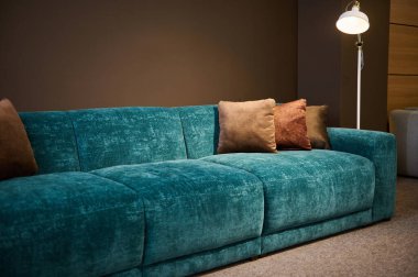 Mobilya mağazasının galerisinde modern şık döşenmiş mobilya sergisi. Turkuaz yumuşak kadife koltuğa odaklan ve bir lamba tarafından aydınlatılmış kahverengi yastıklara.