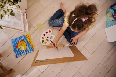 Avrupa 'nın sevimli küçük kızı resim yapmaktan zevk alıyor, ahşap bir sehpada, renkli neşeli çocukların arka planında oturuyor. Sanat sınıfı, yaratıcılık ve eğitim