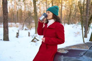 Kıvırcık saçlı esmer, sıcak kış kıyafetleri içinde kadın gezgin, termo bardaktan sıcak çay içiyor, araba yolculuğu sırasında dinleniyor, güzel güneşli bir günde karlı bir ormanda arabanın kaputunda oturuyor.
