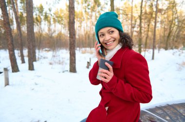 Kıvırcık saçlı esmer kadın sürücü cep telefonuyla eğlenceli sohbetler yapıyor, elinde termometreyle gülümsüyor ve karlı ormanda arabanın kaputunda oturmuş kahve molası veriyor.