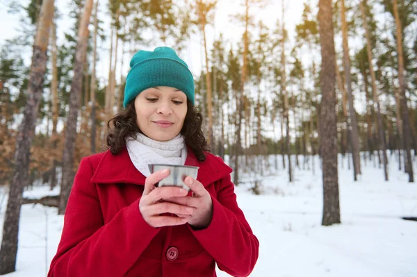 Çekici, çekici, çarpıcı, kırmızı ceketli, yeşil örülü yün şapkalı, elinde termo bardakla genç bir kadın güneşli bir kış gününde, karlı bir havada kahve molası veriyor.