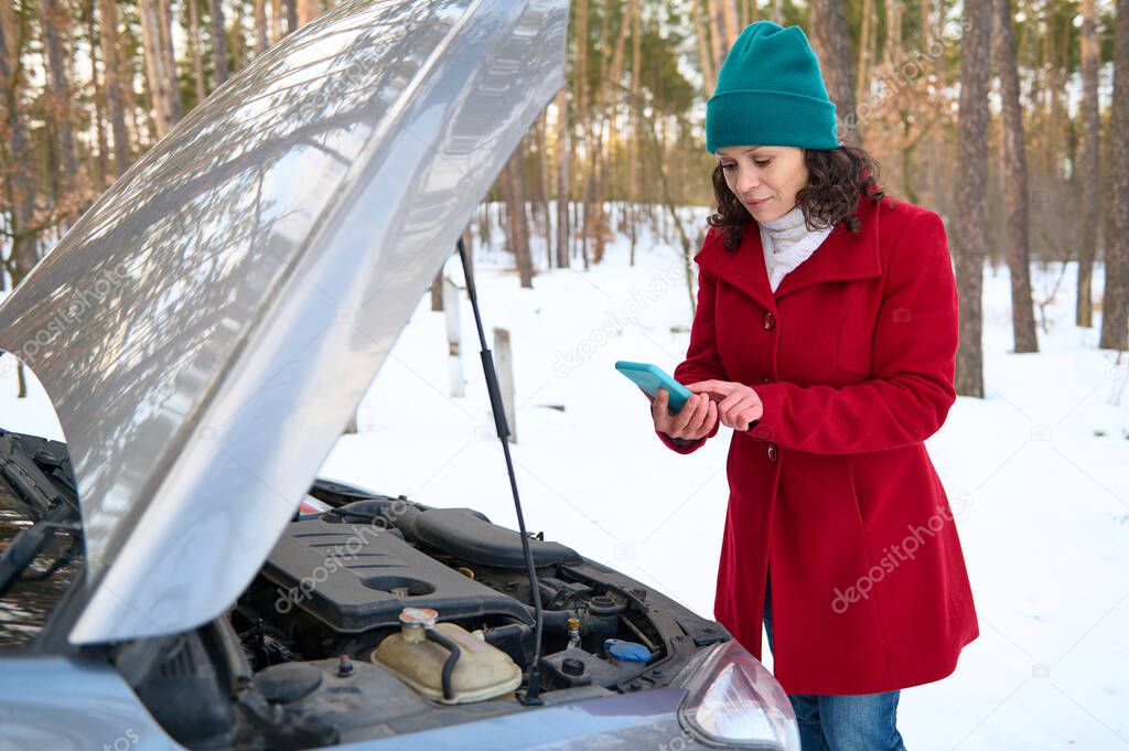 A female driver dials the number of a tow truck or roadside assistance, standing in front of a car with an open hood on a snowy road. Concept of car breakdown during travel and auto insurance