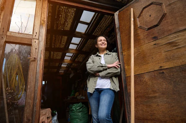 Tepeden tırnağa kendine güveni tam bir kadın bahçıvana bakın, neşeli bir kadın, kollarını kavuşturmuş, tahta kırsal atölyesinde duruyor bahçe aletleriyle ve kameraya bakarak gülümsüyor