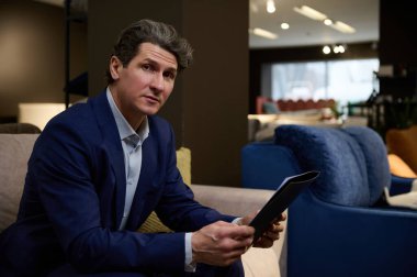 Mobilya dükkanındaki koltukta oturmuş, kendinden emin bir şekilde kameraya bakan yakışıklı, beyaz bir adam.