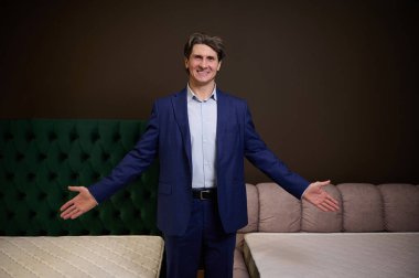 Kendinden emin, orta yaşlı, satış müdürü iş kıyafetleri giyiyor, mobilya mağazasında asistanı ortopedik yatak sunuyor, kameraya bakıp gülümsüyor.