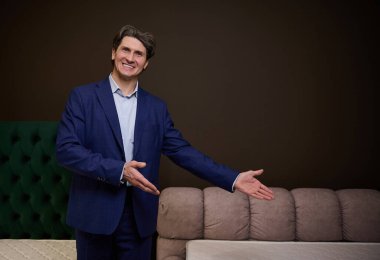 Yakışıklı, orta yaşlı beyaz bir adam, satış müdürü, danışman, şık, yumuşak bir yatağı ve mobilya mağazasındaki ortopedik bir yatağı işaret ediyor. Sağlıklı uyku ve iyi duruş konsepti