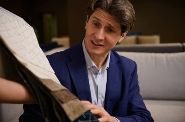 Takım elbiseli yakışıklı, orta yaşlı bir adamın yakın plan çekimi yeni bir kanepe için döşeme malzemesi seçimine şaşırır. Mobilya mağazasındaki bir satış elemanının elinde kumaş örneklerini inceler.