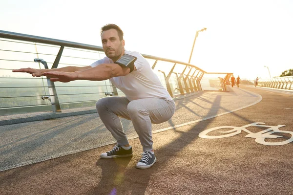 中年アスリート スポーツマンは 夜明けに街の橋の上にスポーツに従事しています 深いスクワットを実行します 体の膝の筋肉 スポーツ エネルギーと動的 健康的なライフスタイルの概念 — ストック写真