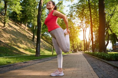 Güneşli bir yaz gününde şehir parkında sabah sporu yaparken bacak kaslarını esneten orta yaşlı bir kadının yan portresi. Spor ve sağlıklı bir yaşam tarzı.