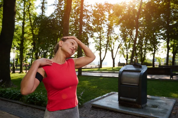 Dar kırmızı tişörtlü, şehir parkında duran, esneme egzersizi yapan ya da sabah koşusundan önce vücudunu ısıtan İspanyol bir kadın.