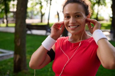 Çekici Afrikalı Amerikalı sporcu, azimli sporcu, parlak kırmızı tişörtlü ve beyaz bileklikli koşucu güneşli yaz günlerinde sabah koşusundan önce kulaklık takarken kameraya gülümsüyor.