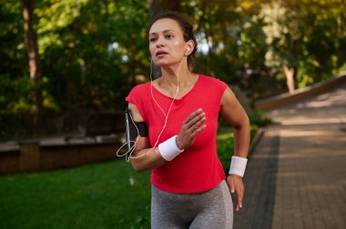 Çekici, bitkin, güçlü kaslı İspanyol sporcu, sporcu, sağlıklı, güzel estetik vücut yapısı olan koşucu şehir parkında koşuyor, güneşli yaz gününde sabah koşusunun keyfini çıkarıyor.