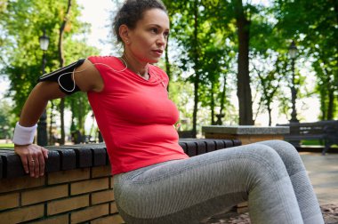 Sportif orta yaşlı Afro-Amerikan kadın dışarıda egzersiz yapıyor. Triseps egzersizleri yapan aktif sporcu bir kadın şehir parkındaki ahşap bankta ellerini yaslıyor.