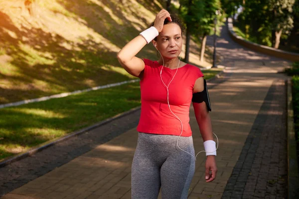 Yorgun bir Afrikalı Amerikalı kadın, kadın koşucu, şehir parkında yürüyor, sabah koşusundan sonra dinleniyor ve güneşli bir yaz gününde kardiyo çalışması yapıyor.