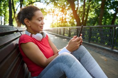 Orta yaşlı Afro-Amerikan atletik kadın spor giyim sektöründe sabahtan akşama kadar ahşap bir bankta dinleniyor cep telefonunu mesaj atmak için kullanıyor, mobil spor uygulamalarını kontrol ediyor.