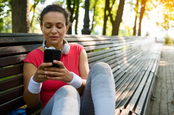 Sportif tarzda gülümseyen Afrikalı Amerikalı atletik kadın ve kulaklıklar güneşli yaz gününde akıllı telefondan dinleme ve mesaj gönderme. Sohbet etmek için telefonu kullanan güzel, neşeli, genç bir sporcu.