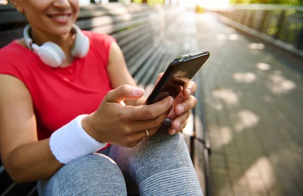Güneşli yaz gününde kulaklık takan ve mesaj atan atletik kadının akıllı telefonuyla ellerine odaklan. Güzel, neşeli genç sporcu kadın antremandan sonra konuşmak için telefonu kullanıyor.