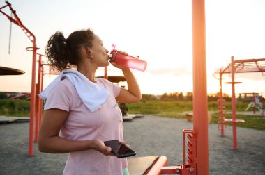 Exhausted middle-aged sportswoman in pink t-shirt with white terry towel drinks water, holds a smartphone and rests after an evening workout at sunset on the sports ground
