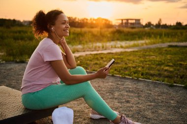Çekici genç Afrikalı Amerikalı bayan atlet, akıllı telefon ve kulaklıklı sporcu akşam antrenmanından sonra gün batımında doğada dinleniyor.