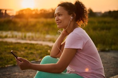 Göz kamaştırıcı güzel Afro-Amerikan kadın kulaklık takıyor, akıllı bir telefon tutuyor ve antrenmandan sonra dinleniyor, gün batımında doğa arka planına karşı sahada oturuyor.