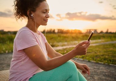 Güzel kulaklıklı İspanyol bayan sporcunun yan portresi, akıllı telefon tutuyor ve güzel günbatımının arka planında kardiyo egzersizlerinden sonra dışarıda dinlenirken mobil fitness uygulamasını kontrol ediyor.
