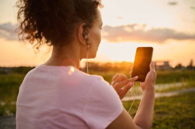 Soft focus on sportswoman hands putting finger on smartphone with blank black screen with copy space for advertising text and mobile fitness app on sunset and nature background